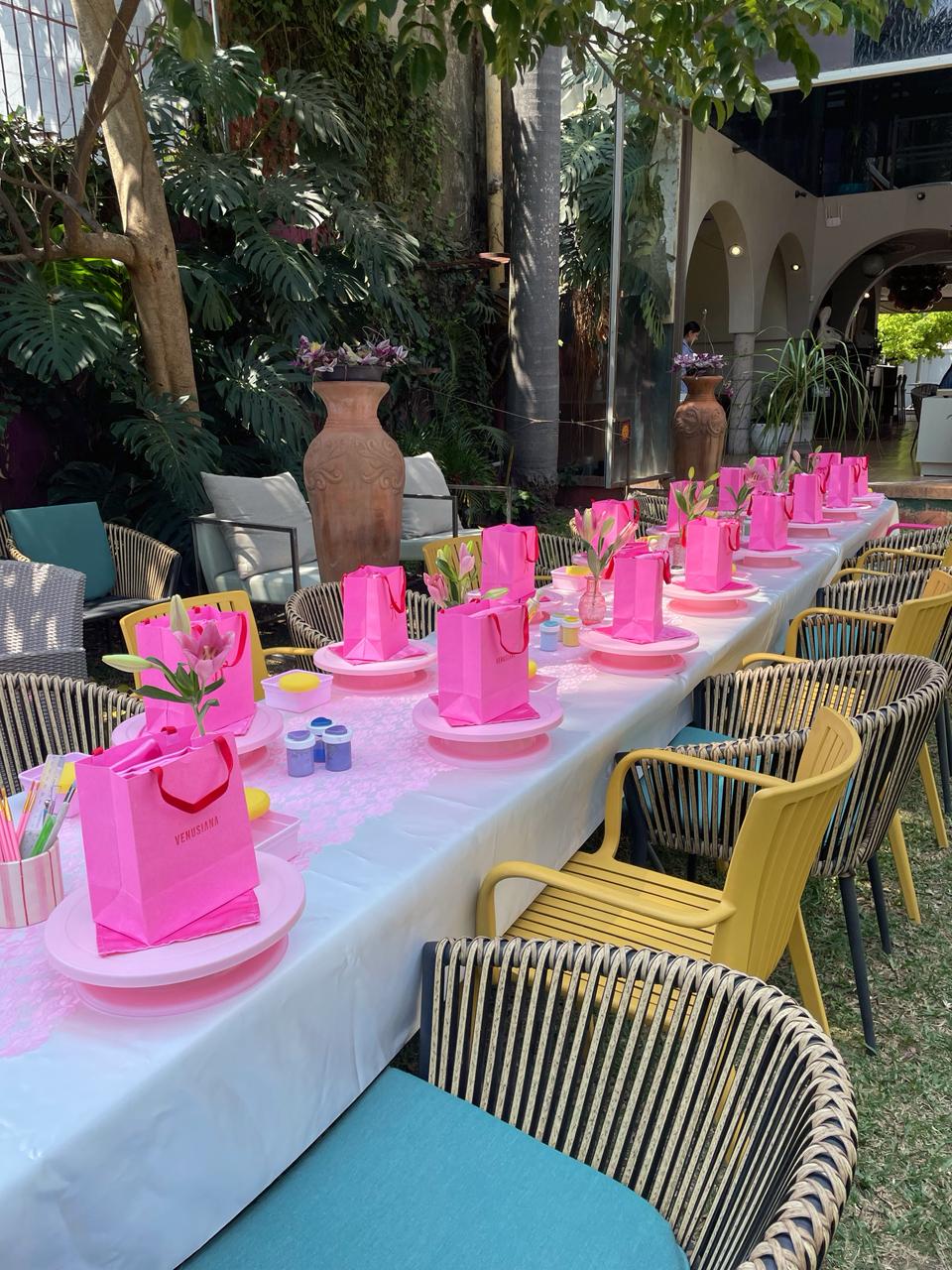 Mesa larga preparada en un jardín con bolsas rosas y bases para modelar